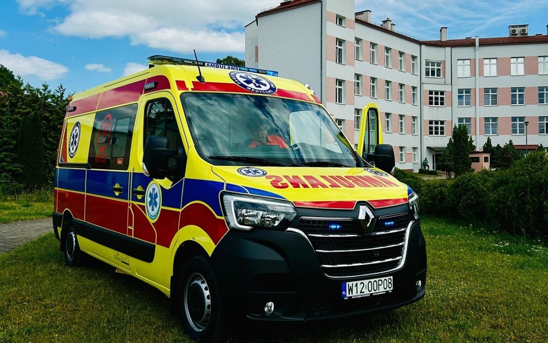 Ambulans na trawniku. Wokół drzewa i krzewy. Za nim budynek szpitala.