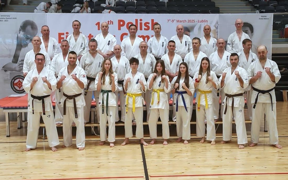 W sali 26 osób stoi obok siebie. Tj. 21 mężczyzn, 4 dziewczyny i chłopak. Są oni ubrani w kimono do karate. Za nimi baner z nazwą zawodów.