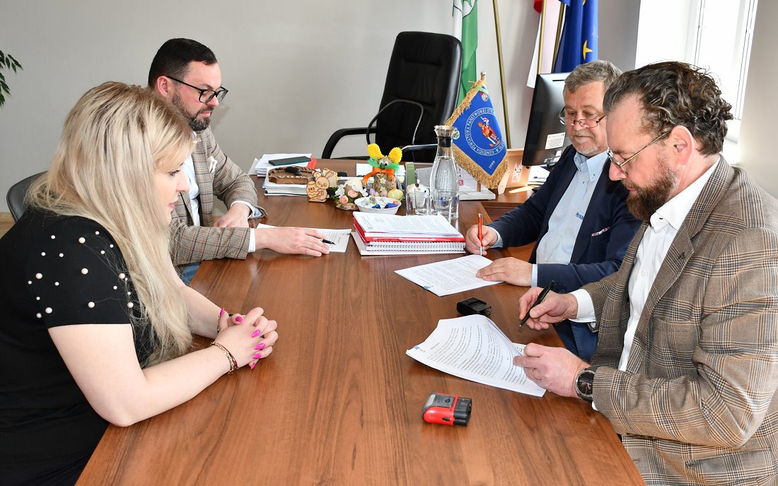 W pomieszczeniu przy stole siedzi 3 mężczyzn i kobieta. Przed mężczyznami leżą kartki i mają oni w dłoni długopisy. Na biurku są pieczątki, dokumenty, różne akcesoria biurowe, karafka z wodą i szklanki.
