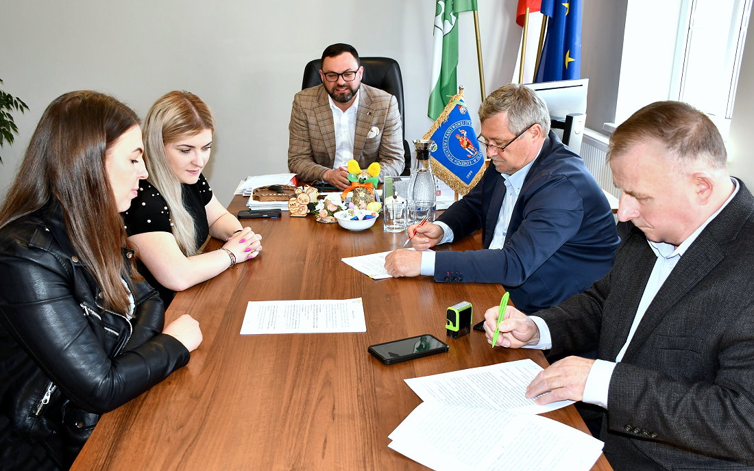 W pomieszczeniu przy stole siedzi 3 mężczyzn i 2 kobiety. Przed 2 mężczyznami leżą kartki i mają oni w dłoni długopisy. Na biurku są pieczątki, dokumenty, telefon, różne akcesoria biurowe, karafka z wodą i szklanki.