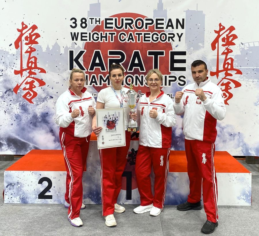 W pomieszczeniu stoją obok siebie 3 kobiety i mężczyzna. Są ubrani na sportowo. Jedna z nich ma puchar i dyplom. Za nimi podium i baner z nazwą zawodów karate po angielsku.